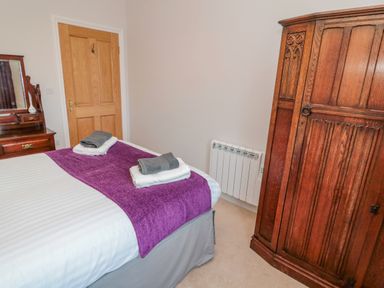 A bedroom with a bed covered with white and purple bedding next to a wooden wardrobe and dresser at High Barns Cottage in Longhorsley near Morpeth