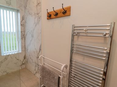 A bathroom corner with marble wall tiles a window with vertical blinds a white towel rack with a gray towel a metal heated towel rail and a wooden wall-mounted coat rack at High Barns Cottage in Longhorsley near Morpeth