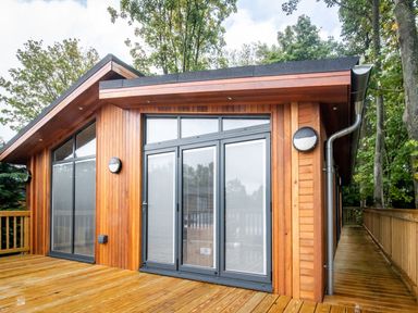A wooden cabin with large glass doors and windows on a wooden deck surrounded by trees at Bluebell Lodge in Gargrave