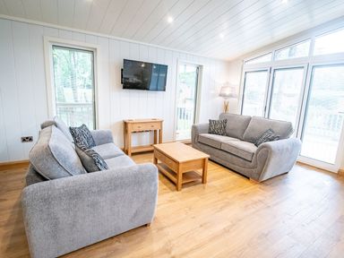 A living room with two grey sofas a wooden coffee table a wooden side table mounted television lamp and large windows at Bluebell Lodge in Gargrave