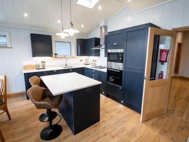 A kitchen with dark cabinets a white countertop stools a wooden floor and a partially open wooden door at Bluebell Lodge in Gargrave