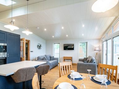 An open-plan living area with a kitchen island and bar stools two grey sofas a wall-mounted TV a round dining table set with white dishes and glasses and large windows at Bluebell Lodge in Gargrave