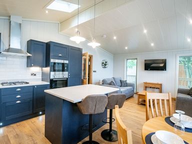 An open kitchen and living area with blue cabinets a kitchen island with stools sofas a wall mounted tv and a dining table at Bluebell Lodge in Gargrave