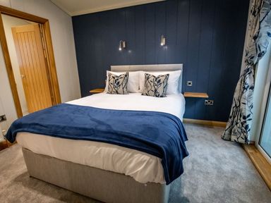 A bedroom with a bed a blue blanket two pillows wooden door and window with floral curtains at Bluebell Lodge in Gargrave