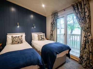 A bedroom with two single beds with blue throws and floral pillows beside a glass door with patterned curtains at Bluebell Lodge in Gargrave
