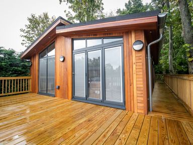 An outdoor wooden deck with railing and glass doors on a wooden cabin surrounded by trees at Bluebell Lodge in Gargrave