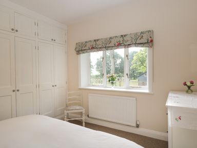 A bedroom with white wardrobes a window with floral curtains and flowers on a window sill and dresser at The Roost in Cheriton near Alresford