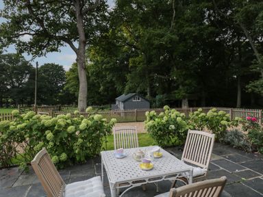 An outdoor patio with a table set with a teapot and cups surrounded by wooden chairs and green shrubs at The Roost in Cheriton near Alresford