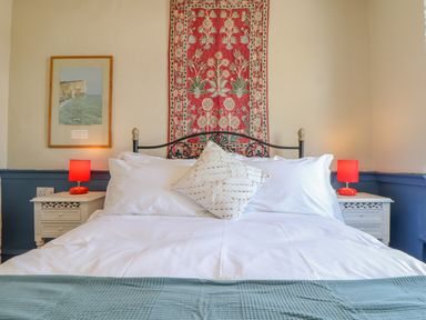 A bedroom with a bed with white linens and a decorative pillow two bedside tables with red lamps a framed picture on the wall and a patterned textile hanging above the headboard at Trewenna Roseland Tregony