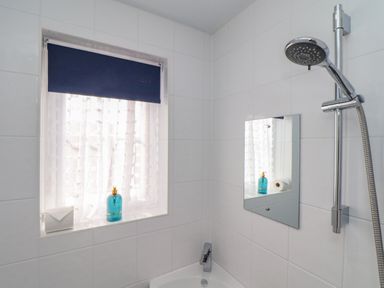 A bathroom with a showerhead a small rectangular mirror and a window with a blue roller blind and white curtain at Trewenna in Roseland Tregony