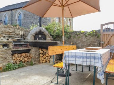 An outdoor patio with a table covered by a tablecloth under an umbrella a built-in stone oven wood logs and wooden benches at Trewenna Roseland Tregony