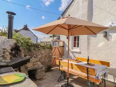 An outdoor patio area with a wooden picnic table and benches under a large beige umbrella next to a stone barbecue grill at Trewenna Roseland Tregony