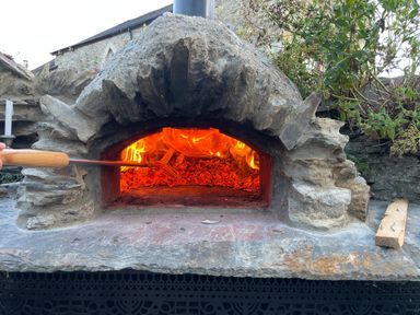 An outdoor stone oven with a fire inside and a wooden handled tool at Trewenna Roseland Tregony