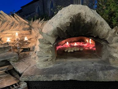 An outdoor stone pizza oven with flames inside and dough balls on the oven floor at Trewenna Roseland Tregony