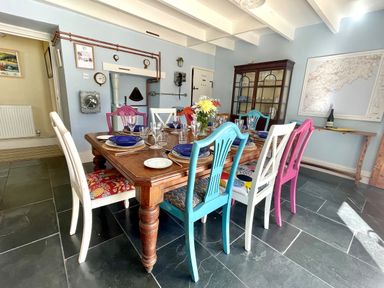 A dining room with a wooden table set for a meal and multicolored chairs at Trewenna in Roseland Tregony