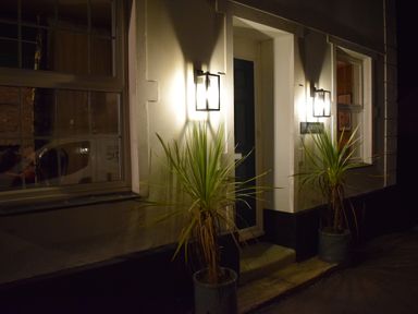 A house entrance with two lit wall lights and potted plants on each side of the door at Trewenna Roseland Tregony