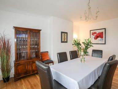 A dining room with a table covered with a white cloth and six chairs a wooden cabinet with glassware and framed pictures on white walls at Awelfryn in Criccieth