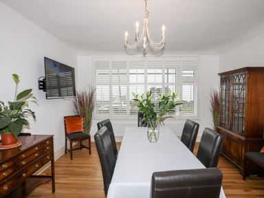 A dining room with a table covered with a white cloth and black chairs around it a vase with flowers on the table a wooden cabinet a TV on the wall and windows with white shutters at Awelfryn in Criccieth