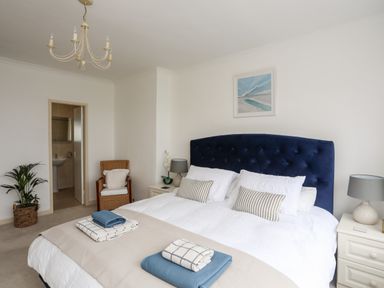 A bedroom with a large bed topped with pillows and folded blankets beside bedside tables with lamps and a wicker chair near a bathroom door at Awelfryn in Criccieth