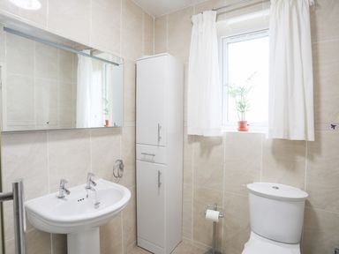 A bathroom with a sink a tall white cabinet a window with white curtains a small plant a toilet and a toilet paper holder at Awelfryn in Criccieth