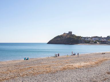 A pebble beach with people near the water and a castle on a hill with buildings in the background at Awelfryn in Criccieth
