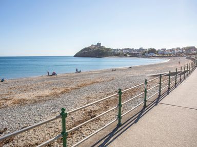 A pebble beach with a walkway and railing and a hill with a castle in the background at Awelfryn Criccieth
