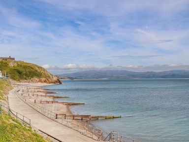 A coastal walkway with railings next to a beach and a grassy hill with ruins at Awelfryn in Criccieth