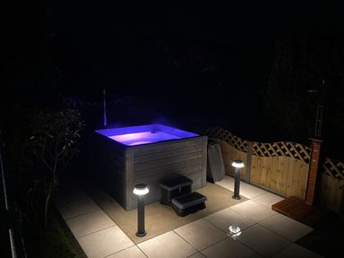 An outdoor hot tub with purple lighting and steps on a tiled patio with lamps and wooden fencing at Awelfryn in Criccieth