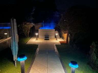 An outdoor night scene with a lit hot tub at the end of a tiled pathway with garden lights and bushes at Awelfryn in Criccieth
