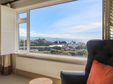 A window with a view of houses trees and water with a chair and cushion by Awelfryn in Criccieth