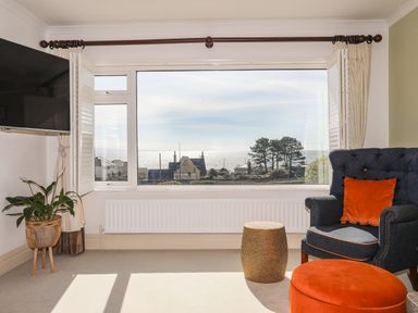A room with a large window showing a sea view a blue armchair with an orange cushion an orange ottoman a plant in a stand and a mounted television at Awelfryn in Criccieth