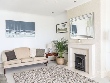 A living room with a beige sofa and cushions a wooden side table with lamp a plant a fireplace with a mirror above and nautical paintings at Awelfryn in Criccieth