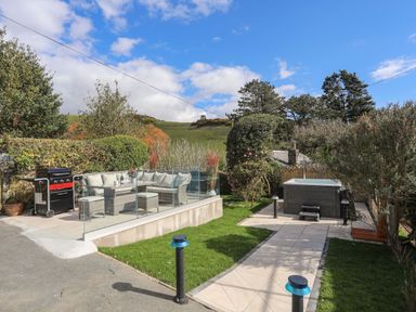 An outdoor patio area with seating and a barbecue grill next to a tiled pathway leading to a hot tub surrounded by greenery at Awelfryn in Criccieth