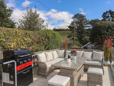 A patio with a gas and coal grill and wicker seating with cushions at Awelfryn in Criccieth