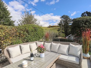 An outdoor patio with wicker seating and a glass table with a bottle, glasses, and a flower pot at Awelfryn in Criccieth