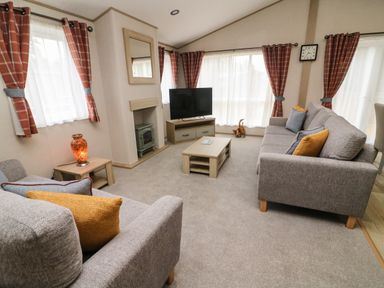 A living room with two grey sofas orange and blue cushions a coffee table tv on a stand and windows with orange curtains at Barneys Retreat in Carnforth
