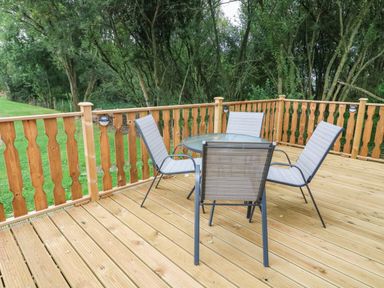 A wooden deck with a round glass table and four chairs surrounded by a wooden railing and trees at Barneys Retreat in Carnforth
