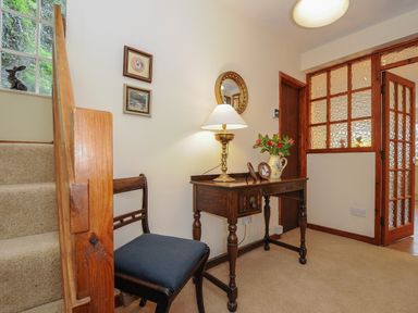 A hallway with a wooden staircase carpeted steps a wooden chair a small wooden table with a brass lamp and vase of flowers two framed pictures on the wall and a glass paneled door at Kirland in St Kew Highway