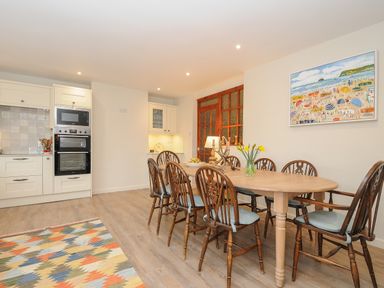 A dining area with a wooden table and eight chairs a kitchen with white cabinetry a colorful patterned rug and a painting on the wall at Kirland in St Kew Highway