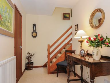 A hallway with wooden stairs a wooden door a radiator a wooden table with a lamp and flowers and a wooden chair at Kirland in St Kew Highway