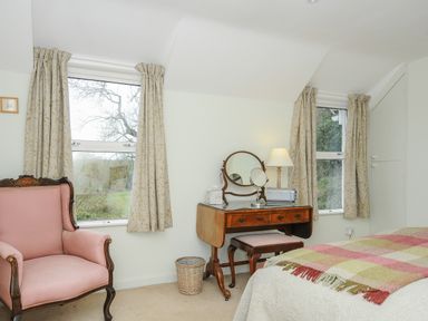 A bedroom with two windows curtains a pink armchair a wooden dressing table with mirrors and a lamp at Kirland in St Kew Highway