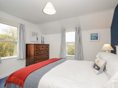 A bedroom with a bed covered in white and red bedding a wooden chest of drawers two windows with striped curtains and a bedside lamp at Kirland in St Kew Highway