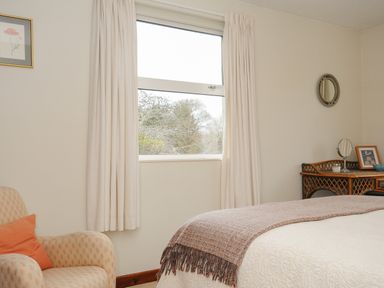 A bedroom with an armchair orange pillow window with curtains a bed with a blanket and a wicker desk with a mirror and framed picture at Kirland in St Kew Highway