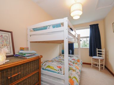 A small bedroom with a white bunk bed a wooden chest of drawers with books a framed picture a window with blue curtains and a white chair at Kirland in St Kew Highway