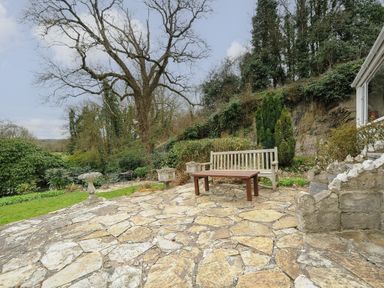 An outdoor stone patio with a wooden bench and table surrounded by trees and shrubs at Kirland in St Kew Highway