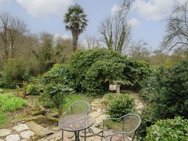 A stone patio with metal chairs and table surrounded by various bushes and trees in a garden at Kirland in St Kew Highway