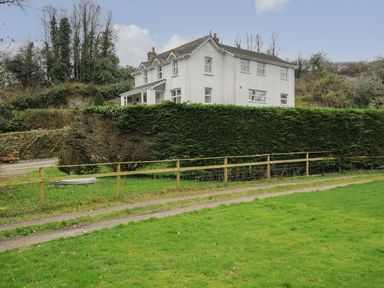 A large white house behind a tall hedge with a wooden fence and grass in the foreground at Kirland in St Kew Highway