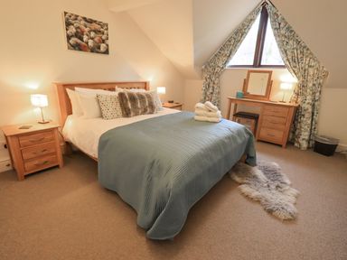 A bedroom with a double bed covered with pillows and a blue quilt next to bedside tables and a dressing table under a triangular window at Beechside in Ambleside