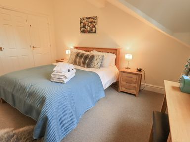 A bedroom with a double bed covered with a blue quilt and folded towels on top two bedside tables with lamps a desk with a chair and a tissue box on it at Beechside in Ambleside