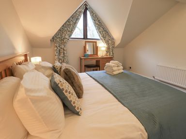 A bedroom with a bed covered in pillows and a folded blanket and towels with a wooden dressing table and triangular window with curtains at Beechside in Ambleside